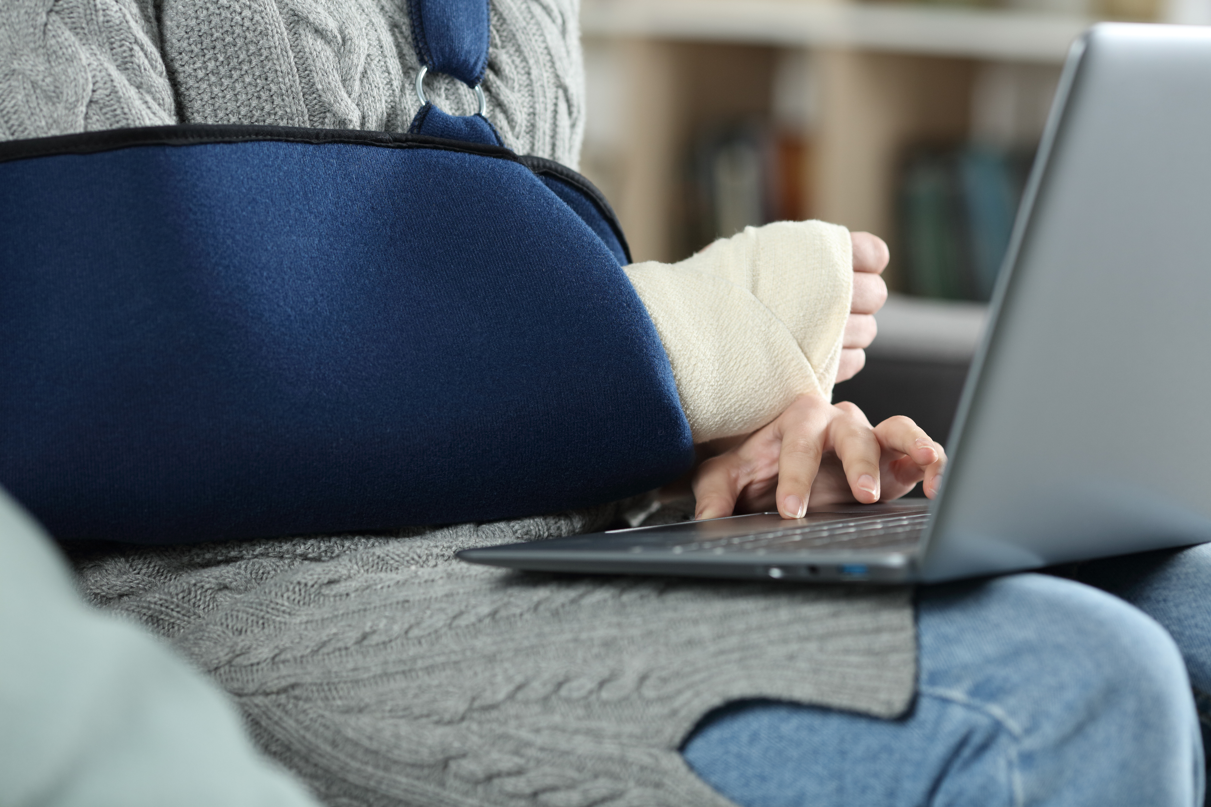 Woman with arm injury wearing a sling while using a laptop at home, representing physical recovery, personal injury challenges, and the complexity of managing medical documentation.